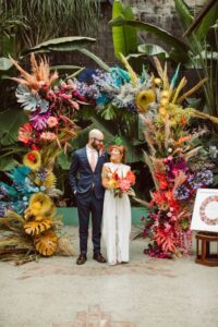 A tropical paradise with tiki torches, exotic floral arrangements, palm leaves, guests wearing tropical prints and leis, and a festive atmosphere with Hawaiian-inspired decor and entertainment